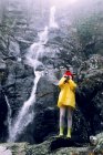 Adolescente anonimo in impermeabile scattare foto sulla macchina fotografica contro il supporto ruvido con schiumosa cascata alla luce del giorno — Foto stock