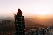 Обратный вид на неузнаваемого туриста в одеяле, стоящего на камне и наблюдающего удивительные пейзажи горной долины в солнечный день — стоковое фото