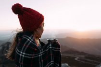 Боковой вид туриста в одеяле, пьющего горячий кофе и стоящего на камне и наблюдающего удивительные пейзажи горной долины в солнечный день — стоковое фото
