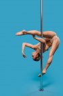 Young flexible barefoot female athlete in bodysuit dancing on pole during workout and looking down on blue background — Stock Photo