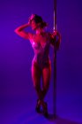 Young female athlete in erotic bodysuit dancing with closed eyes and crossed legs near pole on purple background — Stock Photo