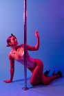Молодая изящная спортсменка в телесном костюме и туфлях на высоких каблуках танцует со скрещенными ногами возле металлического полюса, отводя взгляд на фиолетовый фон — стоковое фото