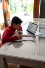 Vista lateral de ángulo alto del colegial inteligente sentado en la mesa con el ordenador portátil y la escritura en el cuaderno mientras hace la tarea solo - foto de stock