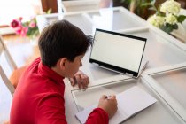 High angle side view of smart schoolboy sitting at table with laptop and writing in notebook while doing homework alone — Stock Photo