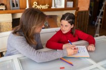 Ângulo alto de mãe ajudando filha com lição de casa enquanto está sentada à mesa com caderno e estudando em casa — Fotografia de Stock