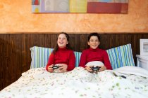 Fröhliche Teenager-Geschwister sitzen auf dem Bett und spielen Videospiele, während sie Gamepads benutzen und das Wochenende zu Hause genießen — Stockfoto