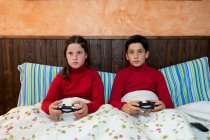 Alegre adolescente irmão e irmã sentados na cama e jogando videogame ao usar gamepads e desfrutar de fim de semana em casa — Fotografia de Stock