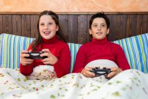 Веселий брат-підліток і сестра сидять на ліжку і грають у відеоігри, використовуючи геймпад і насолоджуючись вихідними вдома — стокове фото