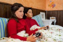 Alegre hermano adolescente y hermana sentados en la cama y jugando videojuegos mientras usa gamepads y disfruta el fin de semana en casa - foto de stock