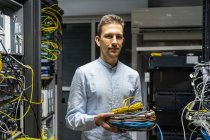 Homem sorridente no fone de ouvido sem fio em pé com cabos na sala do servidor de rede para fornecer Internet e comunicação — Fotografia de Stock