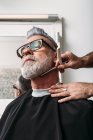 Baixo ângulo de cultura barbeiro masculino irreconhecível barba cinzenta barba de cliente elegante de meia idade em óculos sentado perto do espelho no salão de cabeleireiro — Fotografia de Stock
