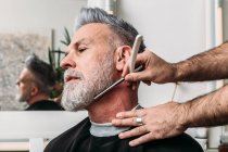 Bajo ángulo de cultivo irreconocible barbero masculino afeitado barba gris de mediana edad cliente elegante en gafas sentado cerca del espejo en el salón de peluquería - foto de stock