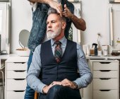 Анонимный парикмахер-стригущий волосы элегантного бородатого мужчины среднего возраста, сидящего на стуле и смотрящего в сторону в современной студии — стоковое фото