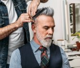 Crop cabelo estilo barbeiro anônimo de elegante bem vestido barbudo cliente masculino de meia idade sentado na cadeira e olhando para longe no estúdio moderno — Fotografia de Stock