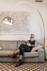 Moderno hombre de negocios de mediana edad navegando por Internet en la tableta mientras está sentado con las piernas cruzadas en el sofá en casa - foto de stock
