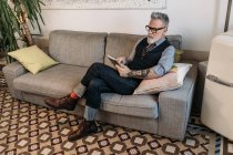Модный бизнесмен средних лет просматривает интернет на планшете, сидя со скрещенными ногами на диване в доме — стоковое фото