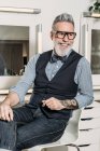 Trendy maturo uomo d'affari hipster in occhiali e vestiti alla moda guardando altrove e sorridendo sulla sedia in casa — Foto stock
