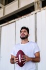Self assured young bearded ethnic male in white t shirt holding pigskin while playing American football on sunny day — Stock Photo