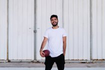 Auto assuré jeune homme ethnique barbu en t-shirt blanc tenant la peau de porc tout en jouant au football américain le jour ensoleillé — Photo de stock
