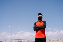 Atleta masculino em pé máscara com os braços cruzados no parque ensolarado antes do treinamento ao ar livre — Fotografia de Stock
