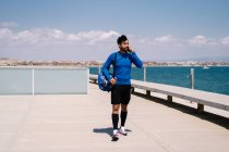 Улыбающийся спортсмен разговаривает по мобильному телефону после тренировки на городской набережной в солнечный день — стоковое фото