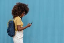 Black woman with afro hair listening to music on mobile in front of a blue wall — Stock Photo