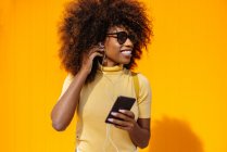 Чорна жінка з афро волоссям слухає музику на мобільному телефоні перед помаранчевою стіною — стокове фото