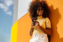 Donna nera con capelli afro che ascolta musica sul cellulare davanti a una parete arancione — Foto stock