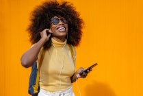 Mujer negra con pelo afro escuchando música en el móvil frente a una pared naranja - foto de stock