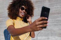 Mujer negra con pelo afro escuchando música en el móvil frente a una pared gris - foto de stock