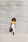 Donna nera con i capelli afro ascoltare musica sul cellulare di fronte a un muro grigio che tiene uno zaino con la mano — Foto stock