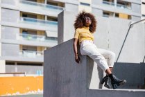 Чорна жінка з кучерявим волоссям сидить на стіні на вулиці і щасливо сміється — стокове фото
