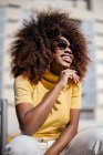 Чорна жінка з кучерявим волоссям сидить на стіні на вулиці і щасливо сміється — стокове фото