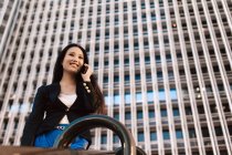 Desde abajo de la mujer empresaria asiática en el estilo casual inteligente de pie en la calle del centro de la ciudad y hablando por teléfono móvil mientras mira hacia otro lado - foto de stock