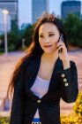 Delighted Asian female entrepreneur standing in street of megapolis in evening and speaking on mobile phone — Stock Photo