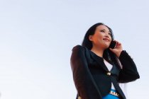 Знизу - жіночий підприємець азіатського стилю, який стоїть на вулиці в центрі міста і розмовляє по мобільному телефону, дивлячись у далечінь. — стокове фото