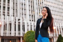 Снизу азиатская предпринимательница в смарт-случайном стиле стоит на улице в центре города и разговаривает по мобильному телефону, глядя в сторону — стоковое фото