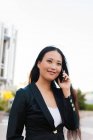 Счастливая азиатская предпринимательница в смарт-случайном стиле стоит на улице в центре города просматривая на мобильный телефон, глядя в сторону — стоковое фото