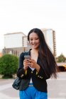 Happy Asian female entrepreneur in smart casual style standing in street of downtown browsing on mobile phone — Stock Photo