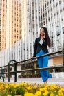 Desde abajo de la mujer empresaria asiática en el estilo casual inteligente de pie en la calle del centro de la ciudad y hablando por teléfono móvil mientras mira hacia otro lado - foto de stock