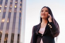 Снизу азиатская предпринимательница в смарт-случайном стиле стоит на улице в центре города и разговаривает по мобильному телефону, глядя в сторону — стоковое фото