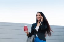 Baixo ângulo de feliz empresária étnica com takeaway café em copo de papel de pé na rua e falando no telefone celular enquanto olha para longe — Fotografia de Stock