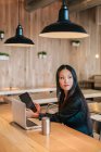 Nachdenkliche ethnische Unternehmerin sitzt mit Tablet und Laptop am Tisch, während sie an einem Projekt arbeitet und wegschaut — Stockfoto