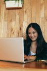 Positive asiatische Unternehmerin sitzt am Tisch im Café und tippt auf Netbook, während sie lächelt und an einem Online-Projekt aus der Ferne arbeitet — Stockfoto