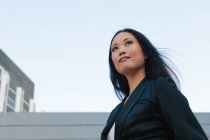 Low angle of determined well dressed Asian female entrepreneur standing with hand in pocket in city street and looking away — Stock Photo