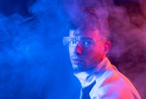 Vista lateral de homem jovem pensativo em roupa elegante e óculos olhando para a câmera enquanto estava em pé no quarto neon fumegante escuro — Fotografia de Stock