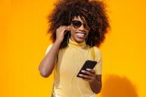 Contenido joven mujer étnica en gafas de sol con peinado afro navegar por Internet en el teléfono celular mientras escucha música sobre fondo amarillo - foto de stock