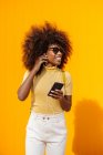 Content young ethnic female in sunglasses with Afro hairstyle browsing internet on cellphone while listening to music on yellow background — Photo de stock