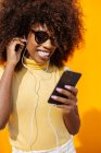 Зміст молодої етнічної жінки в сонцезахисних окулярах з африканською зачіскою переглядає інтернет на мобільному телефоні, слухаючи музику на жовтому тлі. — стокове фото