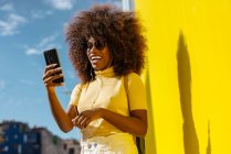 Contenuto giovane femmina etnica in occhiali da sole con acconciatura afro navigare in internet sul cellulare durante l'ascolto di musica su sfondo giallo — Foto stock